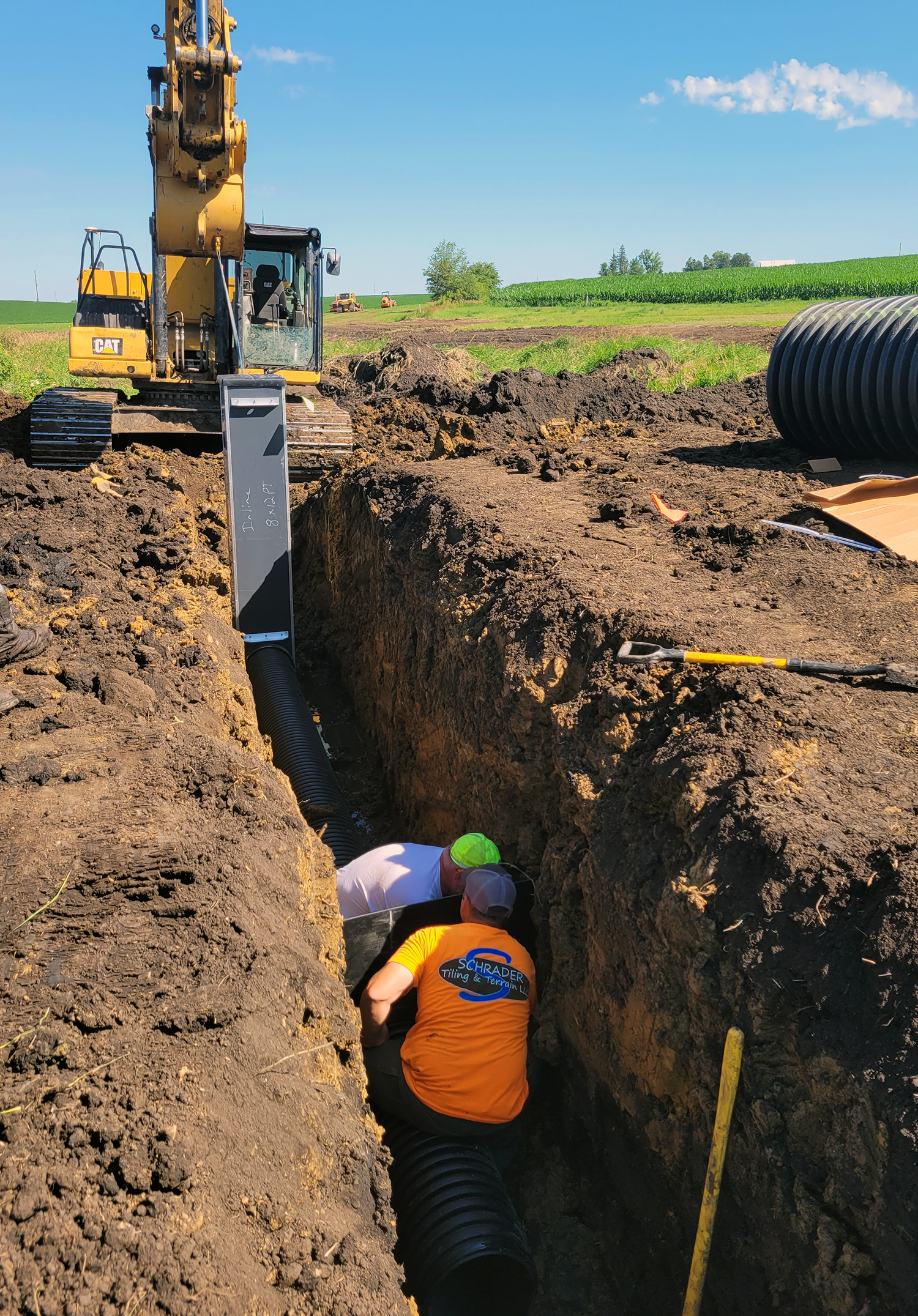Two workers set a large pipe into a deep, earthen trench. The hose is connected to a saturated buffer control unit aboveground, with an excavator in the background.